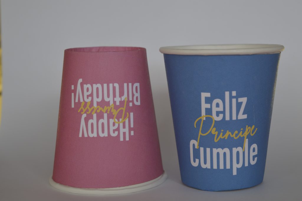 Par de vasos con fondo de color, vaso rojo boca abajo con letreo de happy birthday princess y vaso azul con letrero de feliz cumple principe