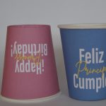 Par de vasos con fondo de color, vaso rojo boca abajo con letreo de happy birthday princess y vaso azul con letrero de feliz cumple principe
