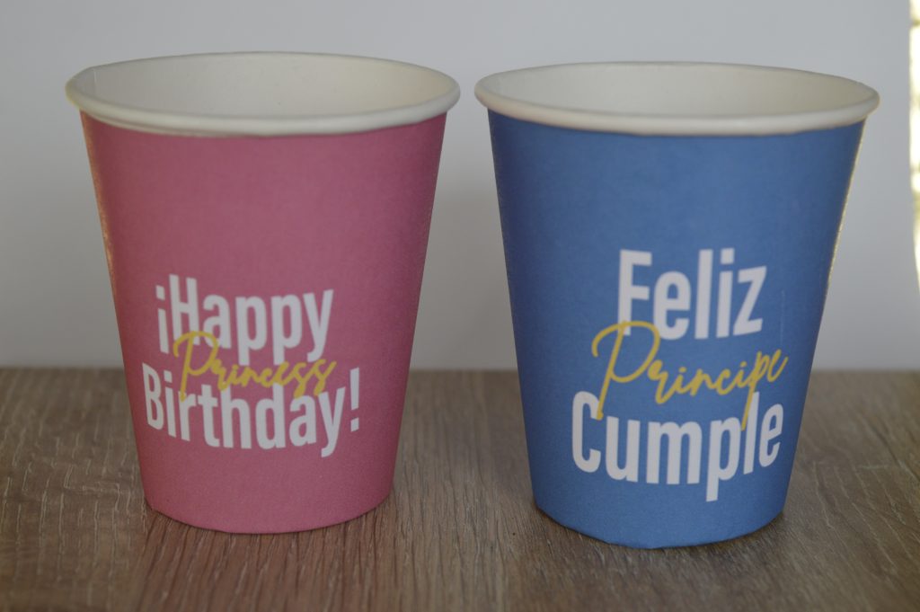 Par de vasos con fondo de color, vaso rojo con letreo de happy birthday princess y vaso azul con letrero de feliz cumple principe sobre una superficie de madera